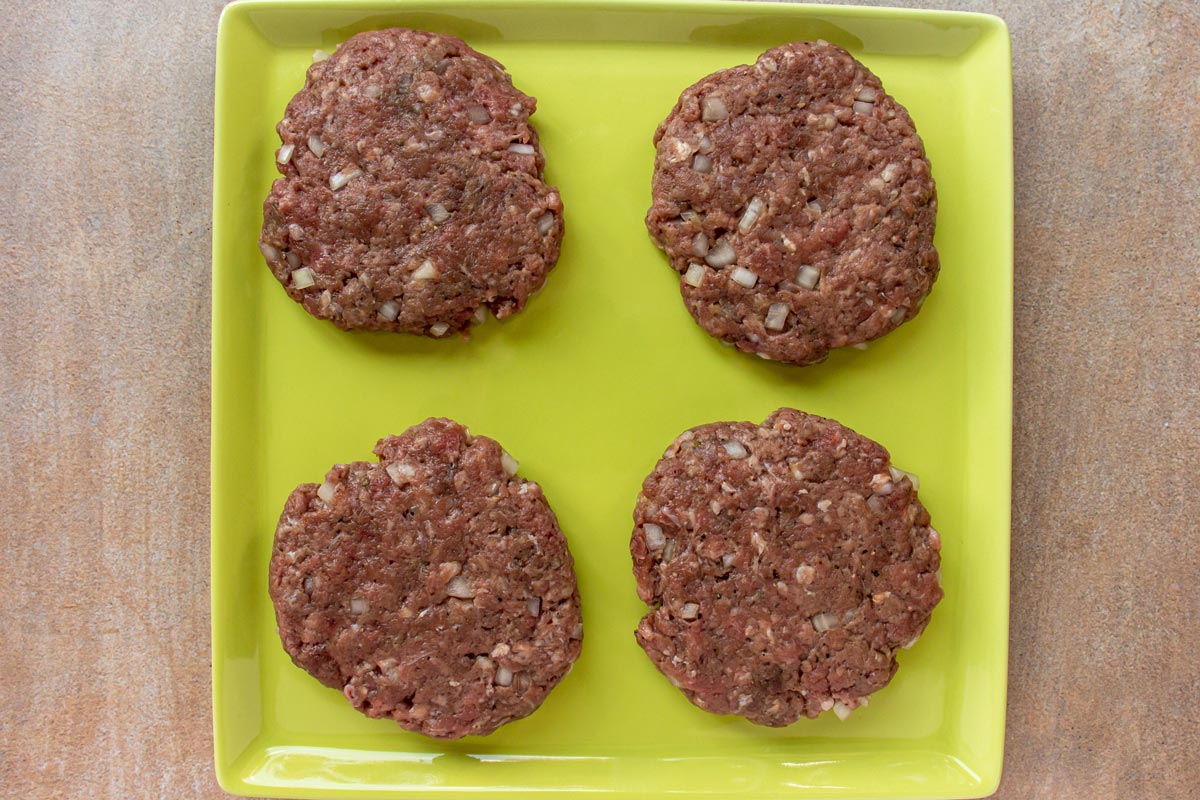 Four uncooked beef burger patties on a light green square plate.