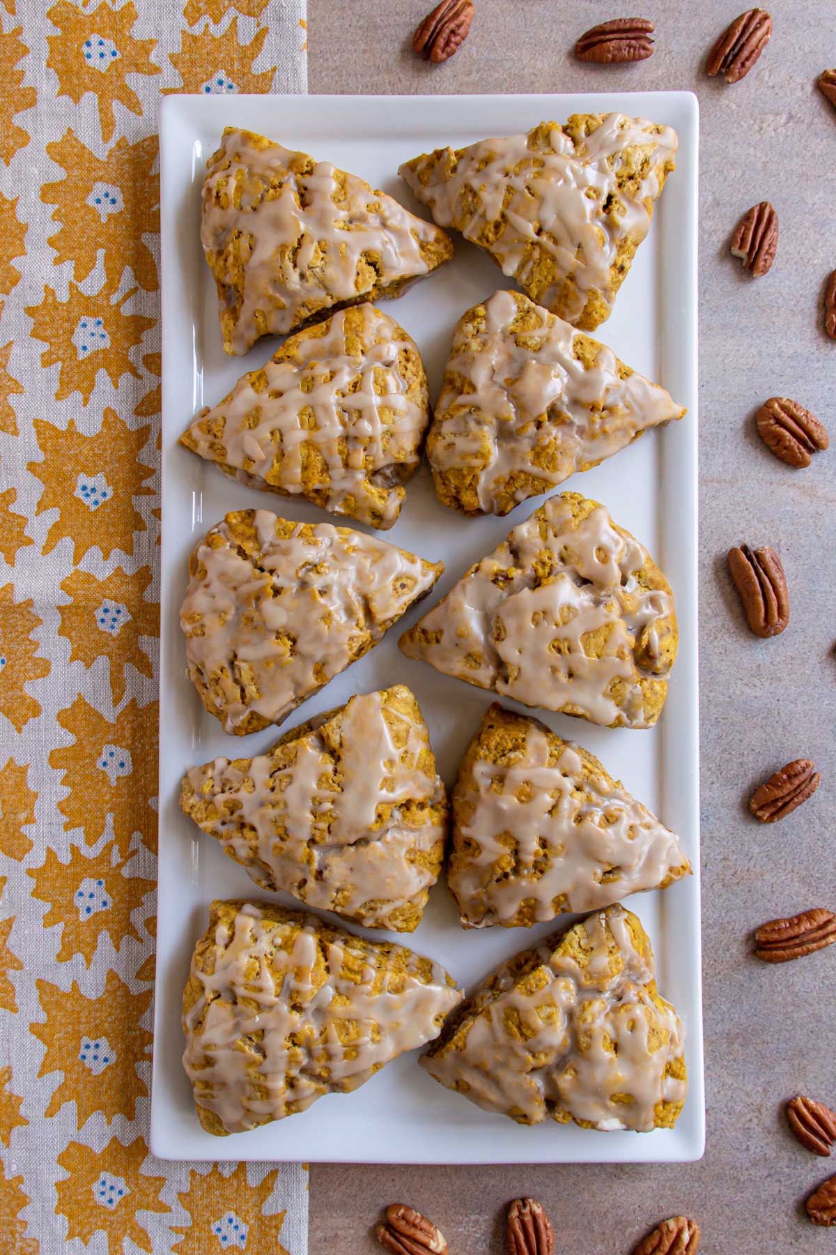 Glazed pumpkin scones on a rectangular plate with pecans and a tea towel on either side.