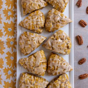 Glazed pumpkin scones on a rectangular plate with pecans and a tea towel on either side.