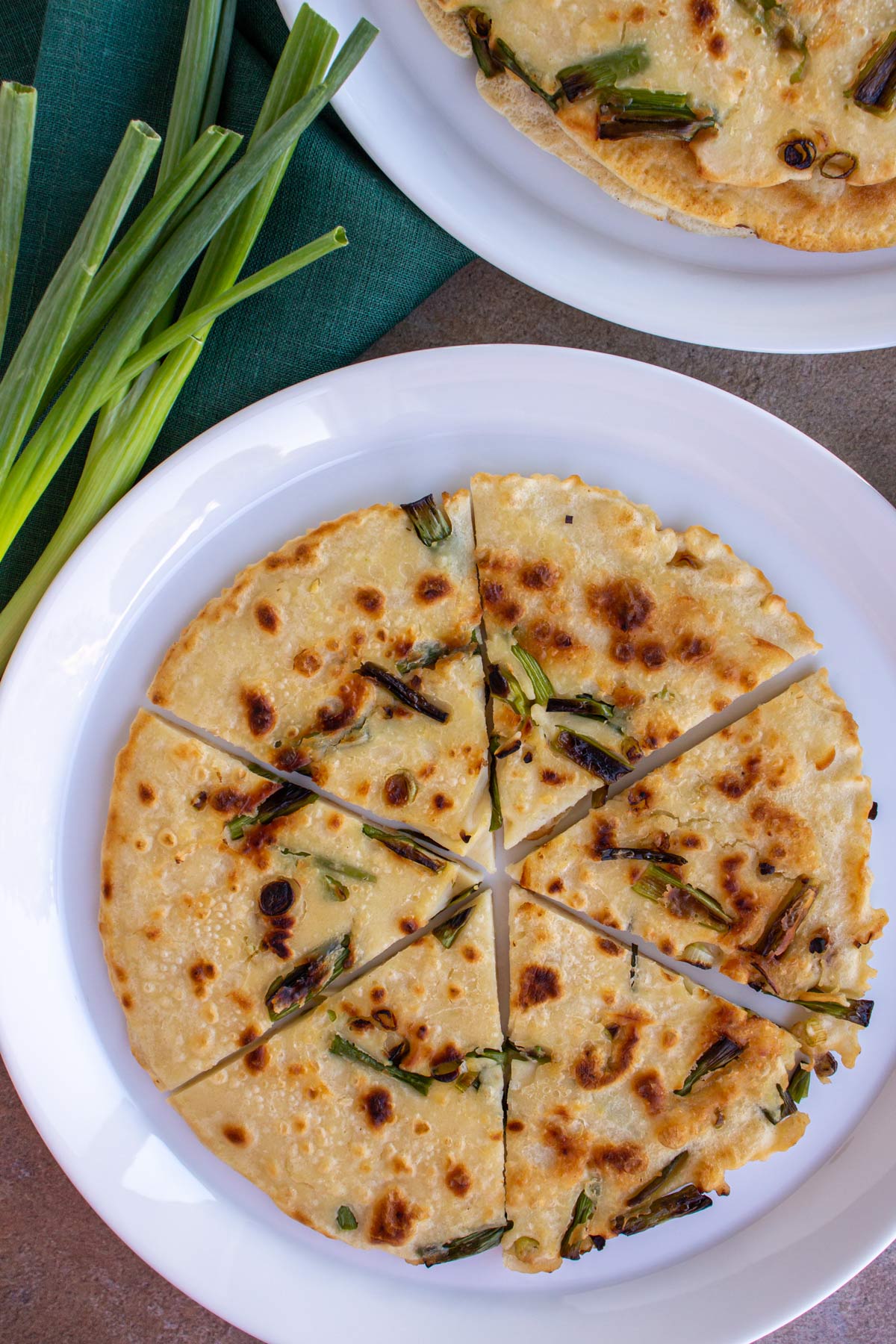 A scallion pancake cut into wedges next to a green linen napkin topped with scallions.
