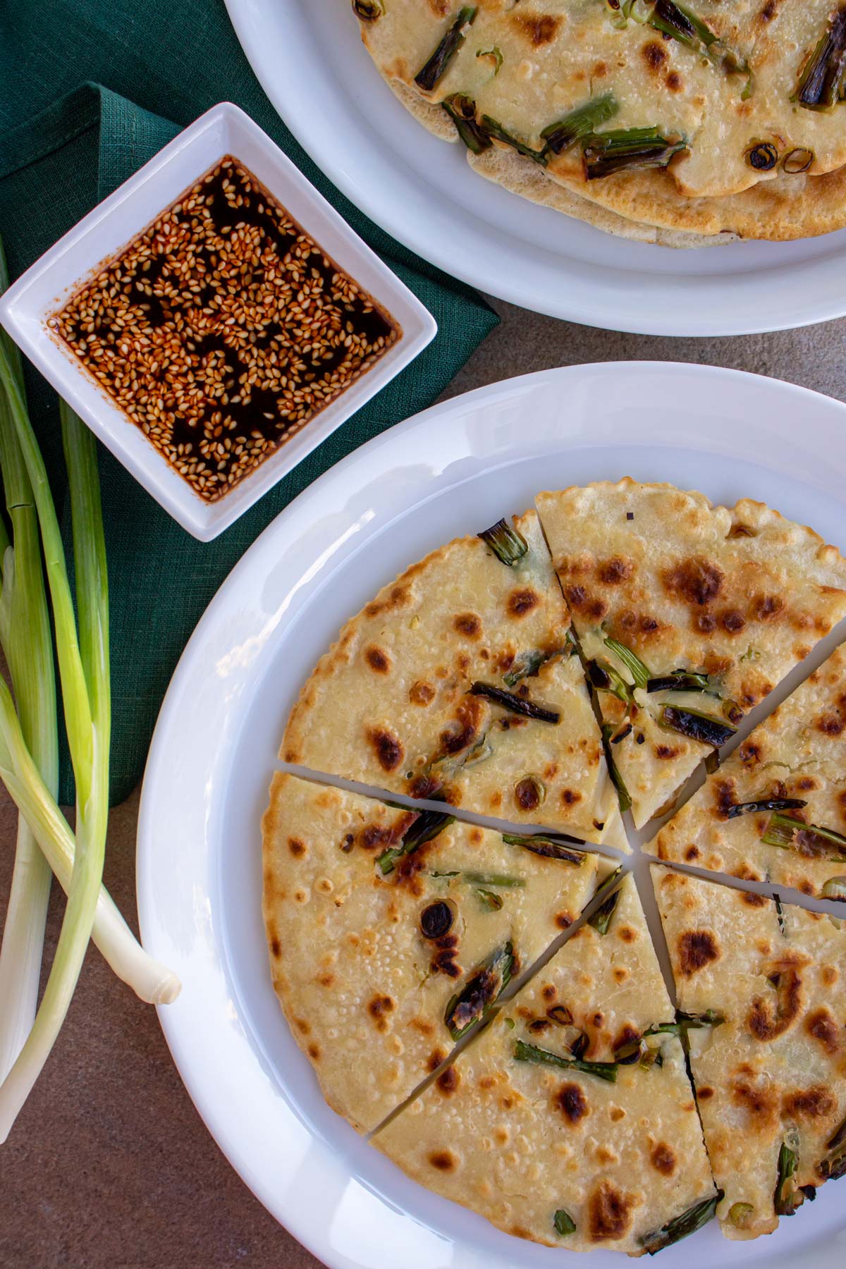 Korean scallion pancakes on white plates with scallions and dipping sauce on the side.