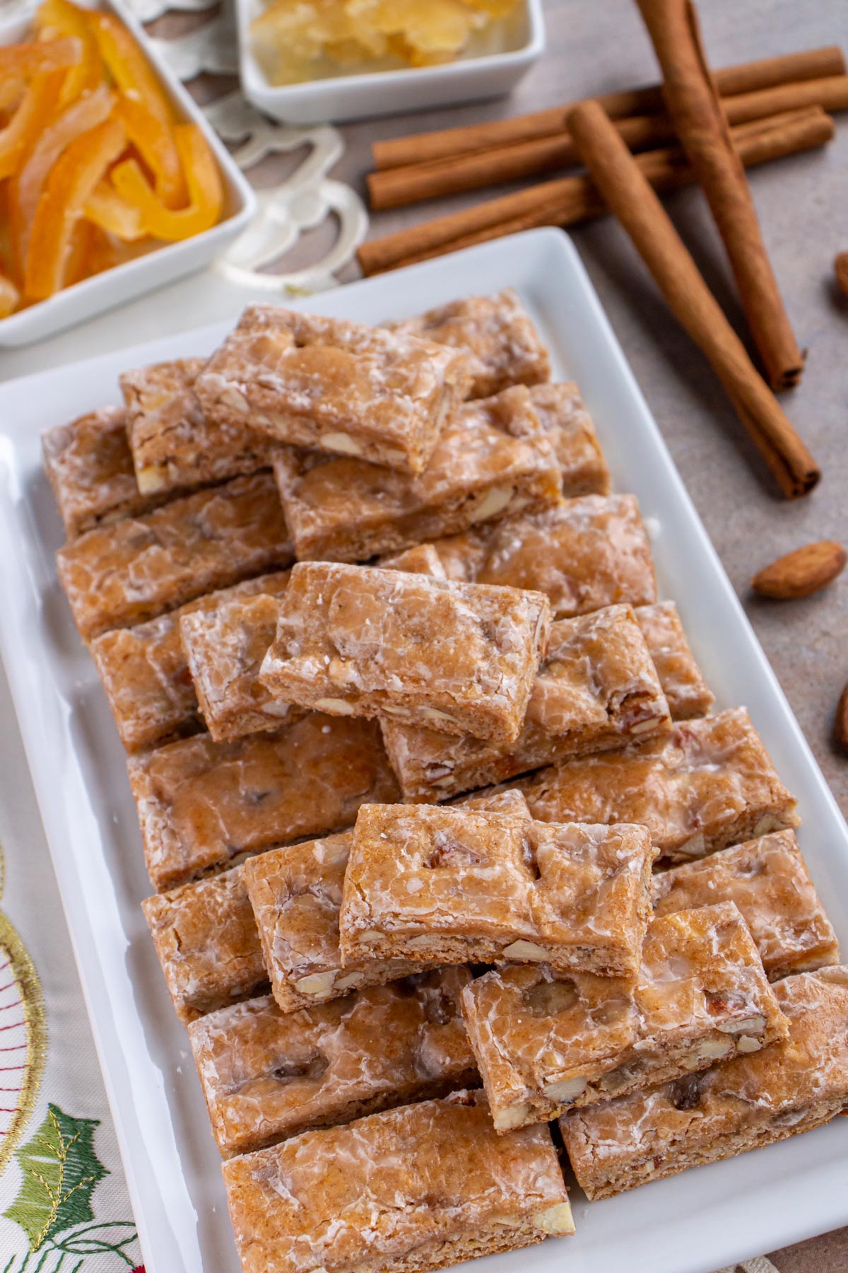 Glazed rectangular cookie bars on a white rectangular plate with candied citrus peel and cinnamon sticks.