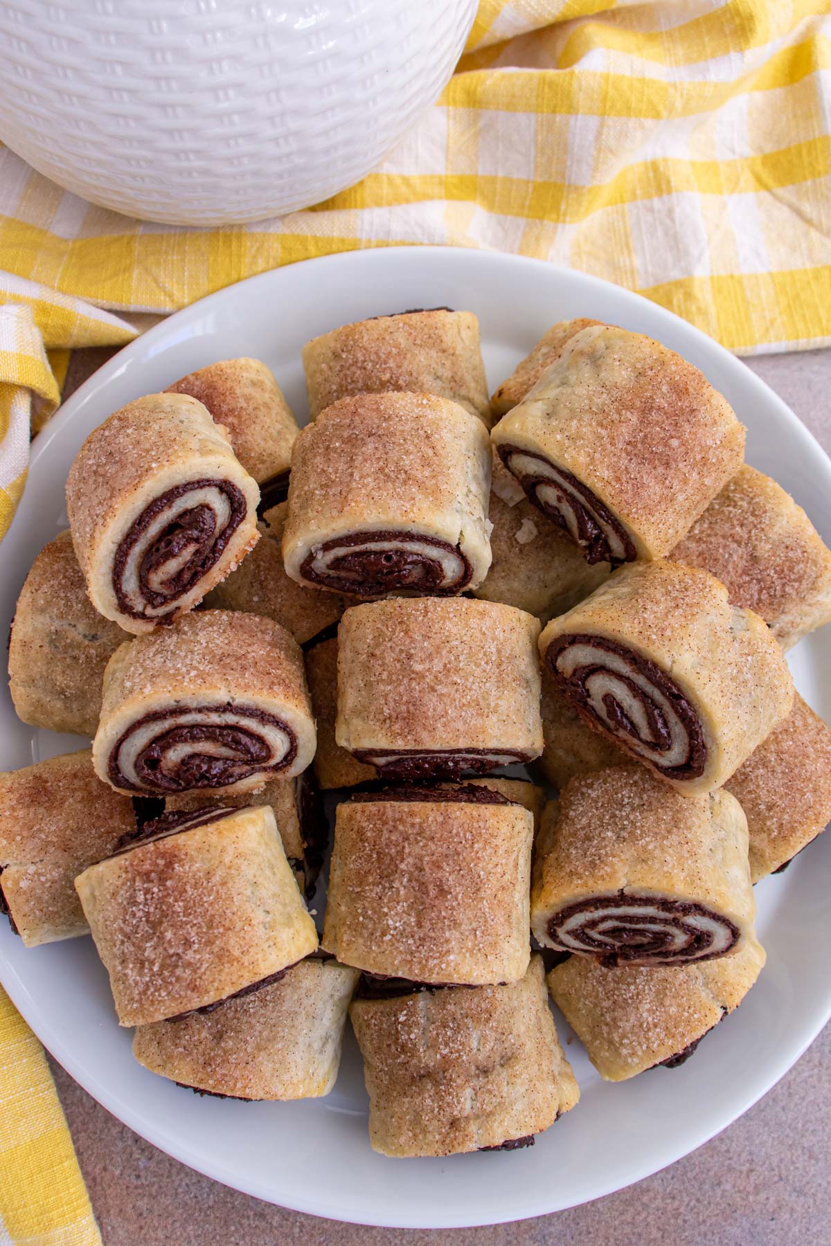 A round platter of chocolate rugelach pastries alongside a yellow gingham towel.