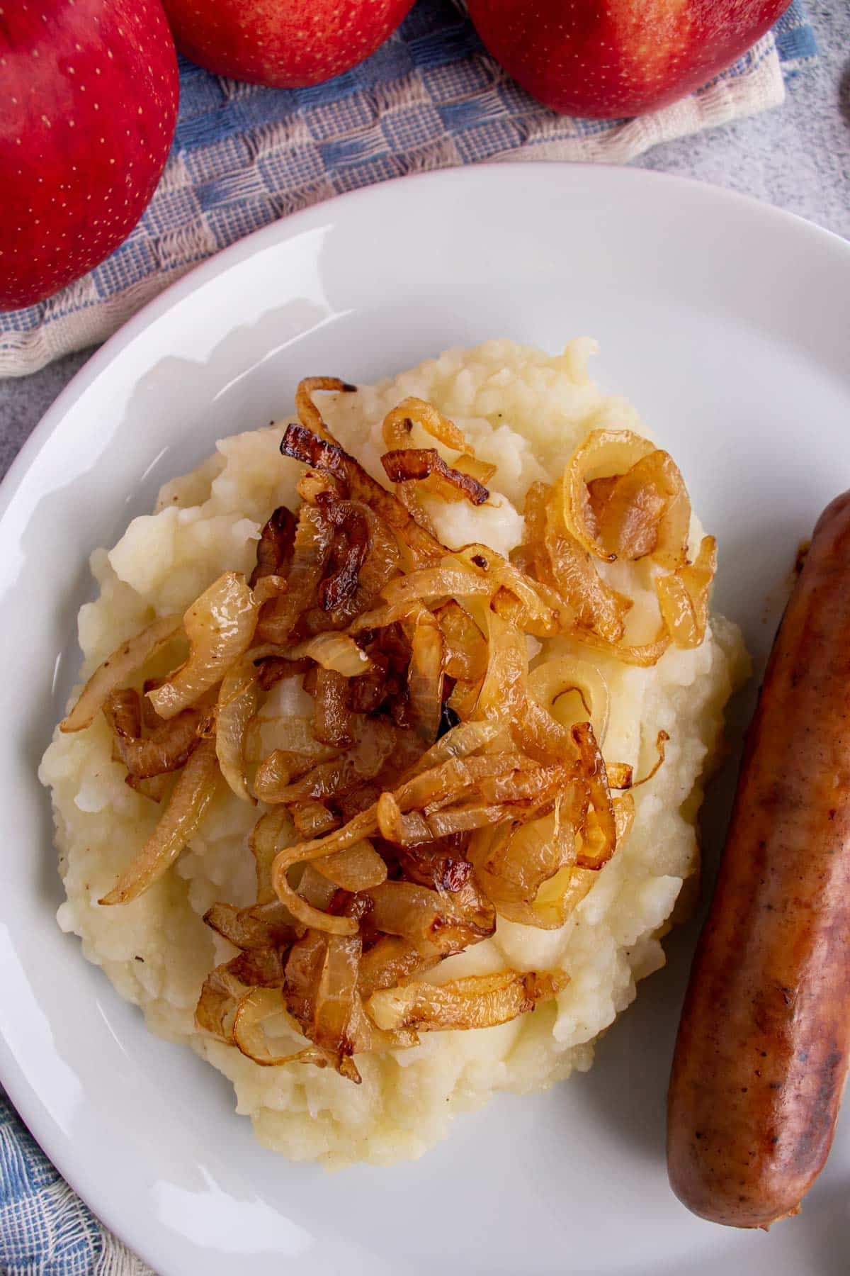 Mashed potatoes and apples topped with fried onions with a sausage on a white plate.