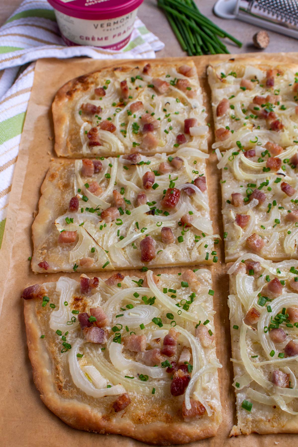 A rectangular flatbread pizza with onions, bacon, and chives cut into 6 squares on brown parchment.