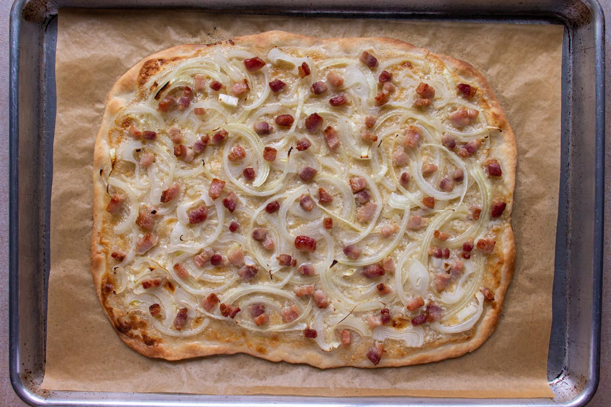 A rectangular baked tarte flambée flatbread pizza with onions and bacon and crispy golden edges.