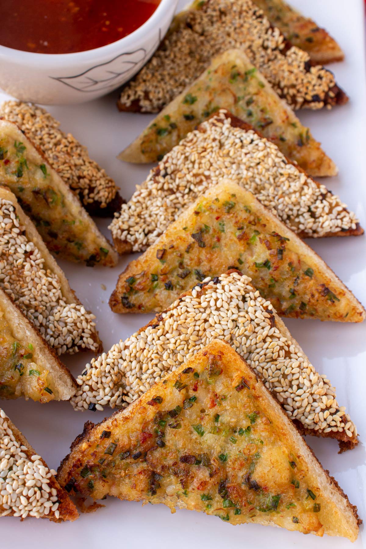Closeup of triangular fried shrimp toasts, half topped with sesame seeds, on a white platter.