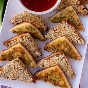 Triangular shrimp toasts, half topped with sesame seeds, arranged in 2 rows on a white platter.