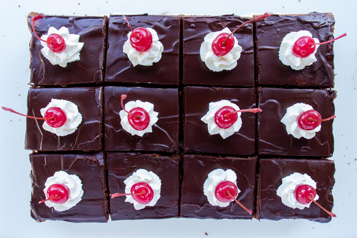 Twelve chocolate ganache topped cake squares garnished with whipped cream rosettes and cherries.
