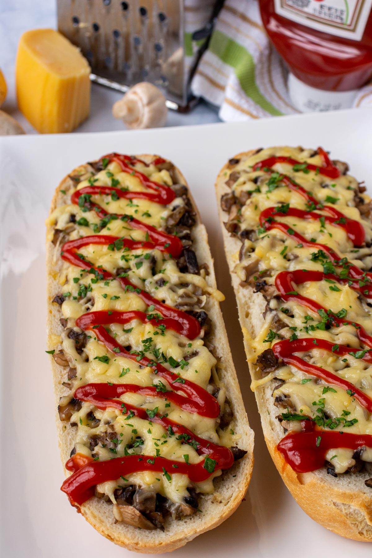 Open-faced baguette sandwiches with mushrooms, melted cheese, and drizzled ketchup on a white plate.