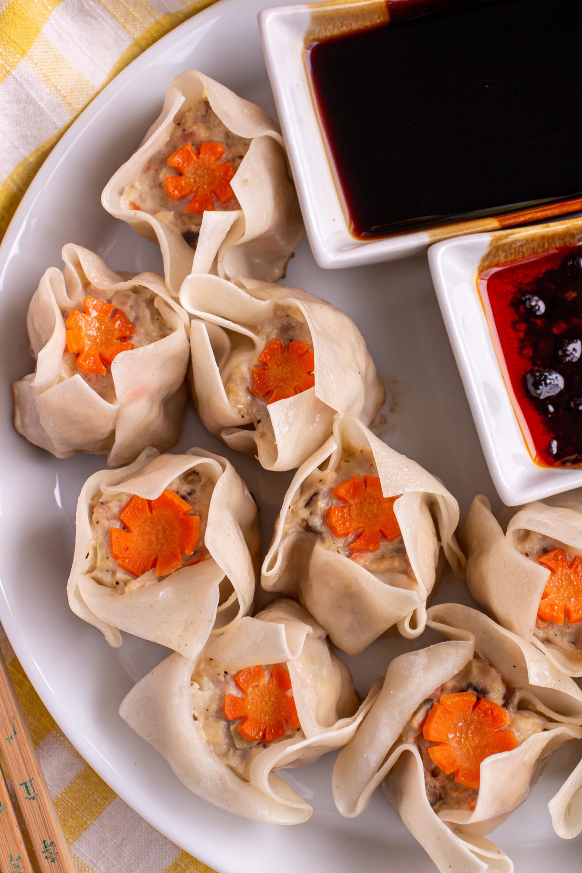 Chicken shumai dumplings topped with carrot garnishes on a white plate with dipping sauce bowls.