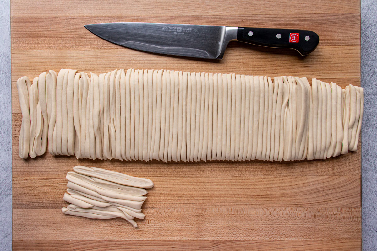 A long strip of dough on a wooden board cut into udon noodles with a knife.