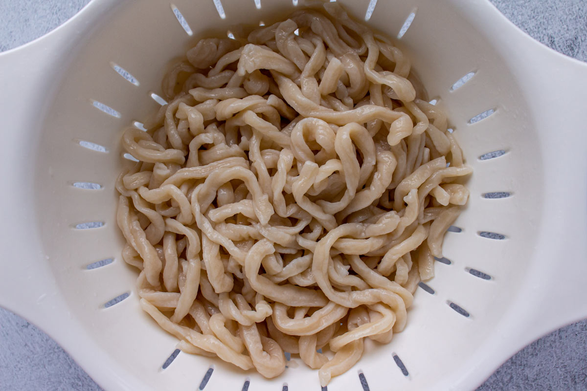 Boiled homemade udon noodles draining in a white colander.
