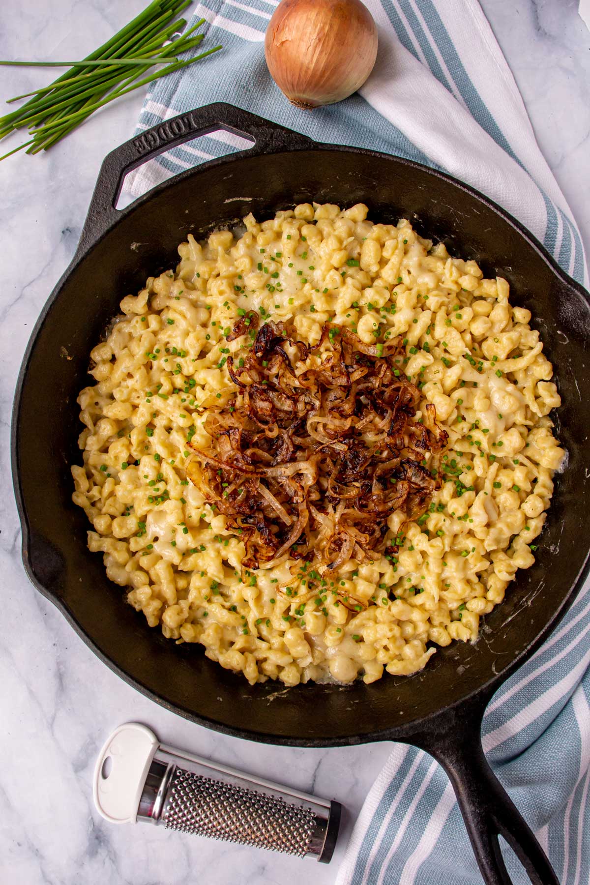 A cast iron skillet of spaetzle topped with fried onions and chives on a marble background.