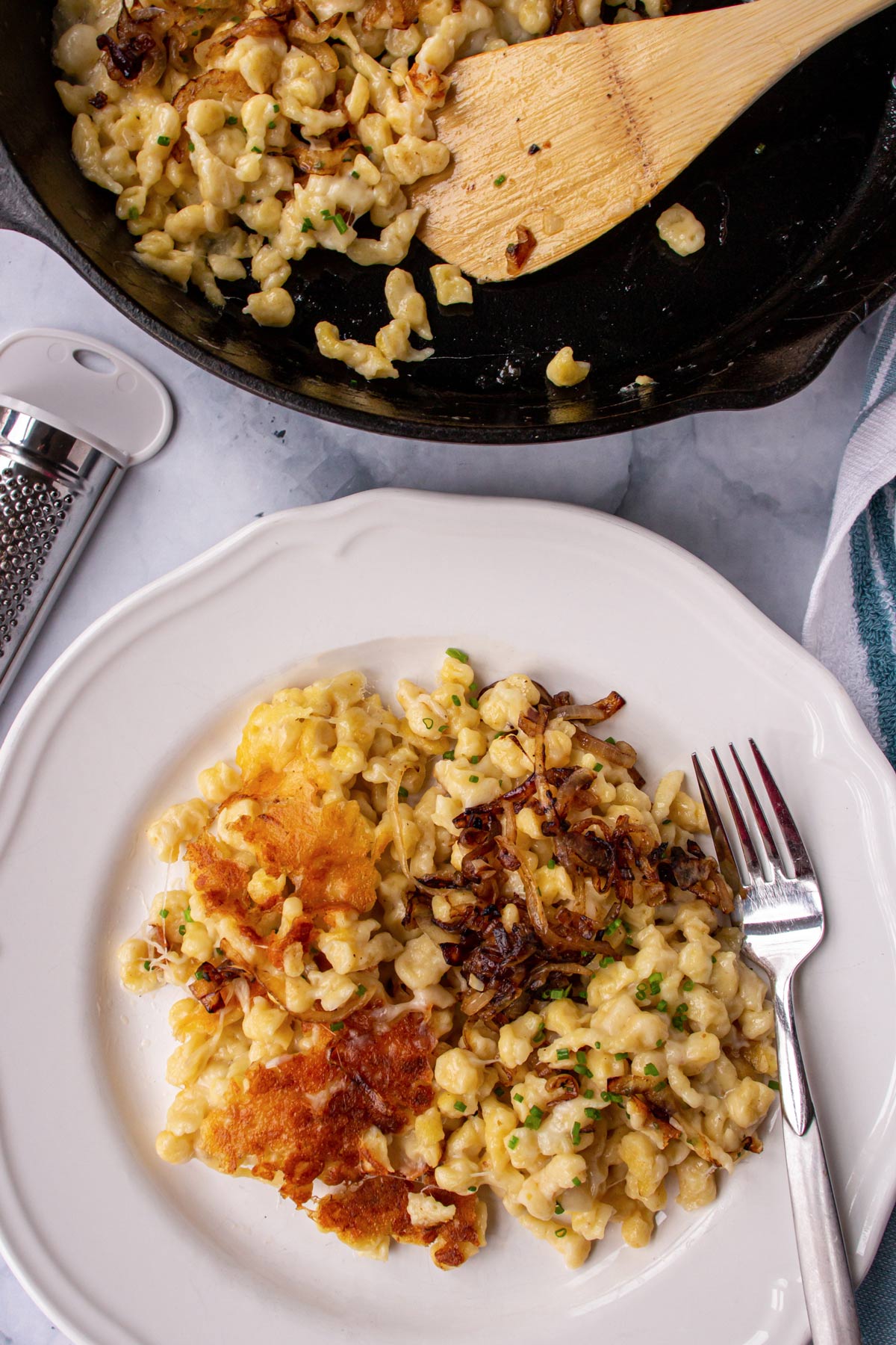 A skillet of kasnocken next to a plate of the cheesy spaetzle dumplings with a fork.