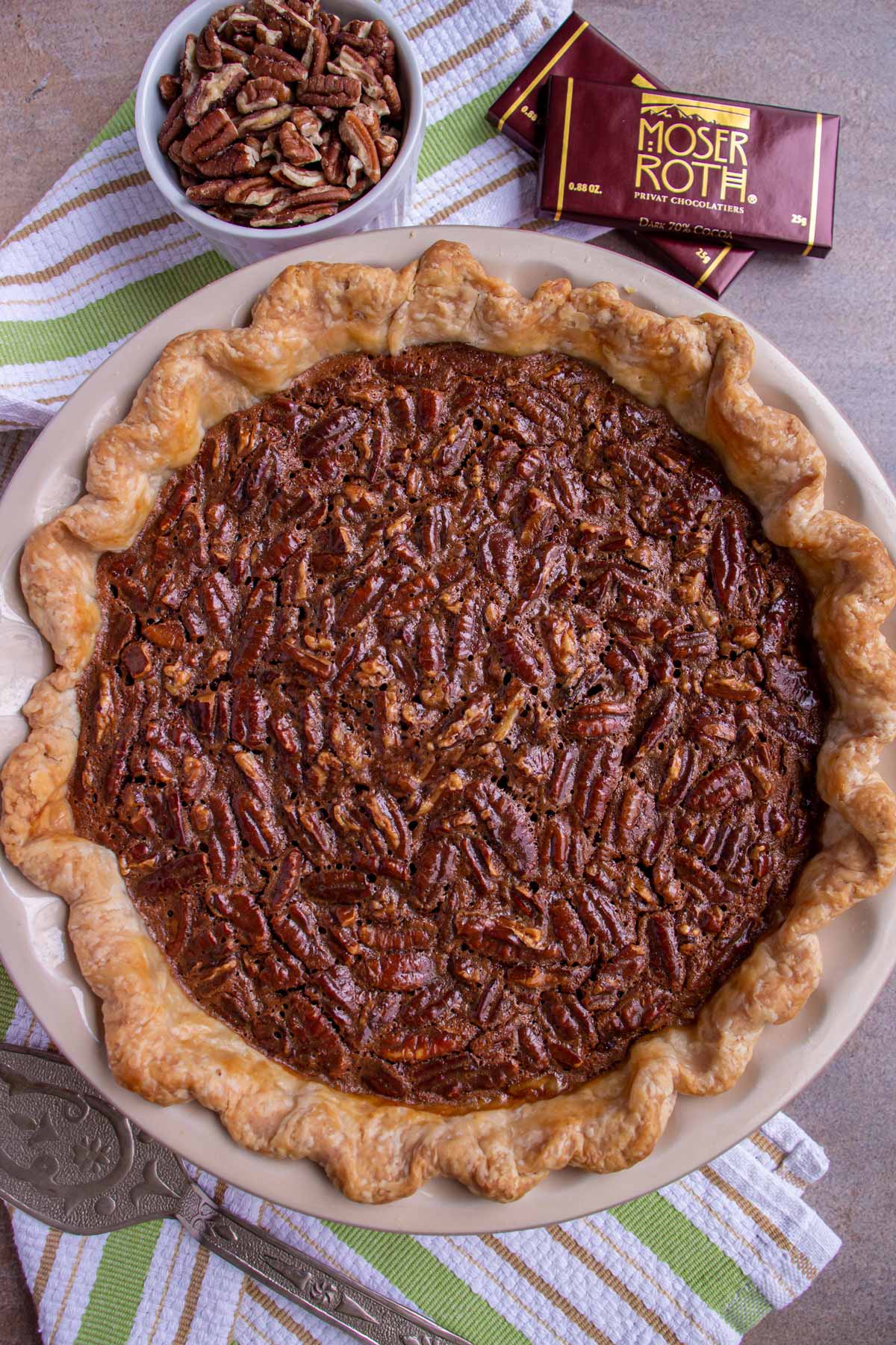 A chocolate pecan pie on a striped towel with pecans and chocolate bars next to it.