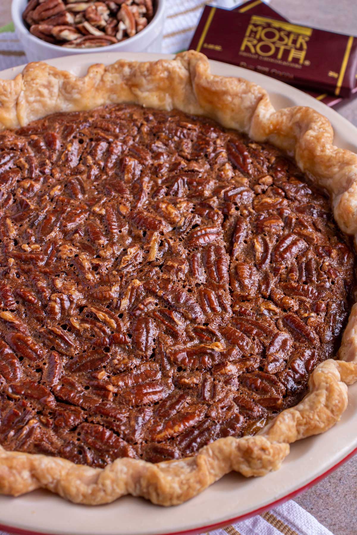 A pecan pie in a ceramic pie dish with pecans and chocolate bars in the background.