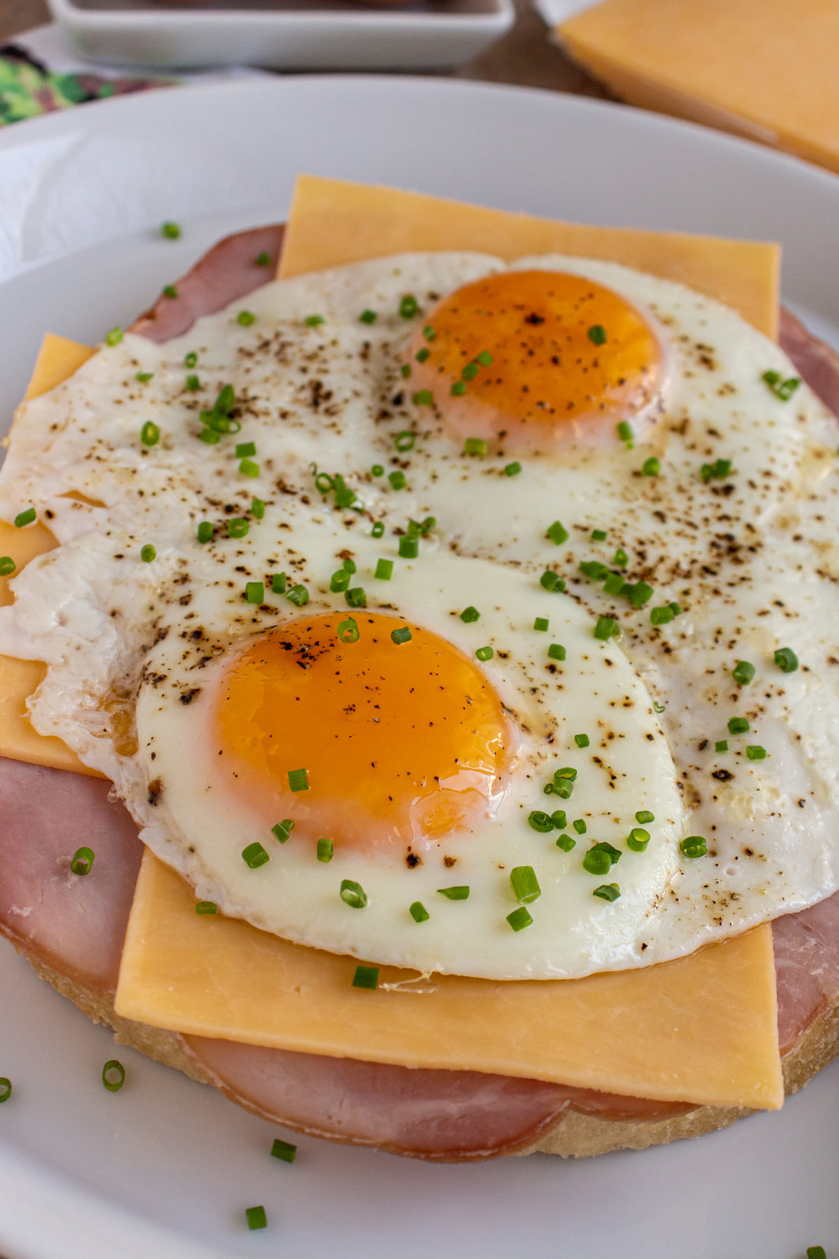 Closeup of sliced bread topped with ham, cheese, fried eggs, and chives on a white plate.