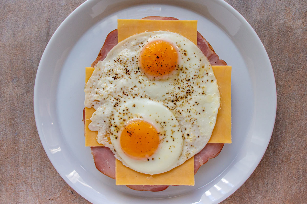 An open-faced sandwich with ham, cheese, and fried eggs on a white plate.