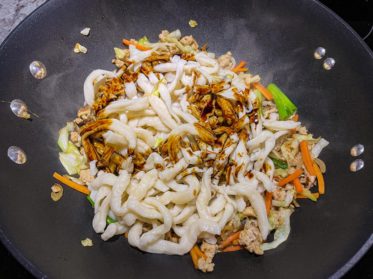 Boiled udon noodles and dark brown sauce added to a mixture in a wok.