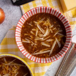 Two colorful bowls of soup topped with grated cheese on a yellow gingham towel.