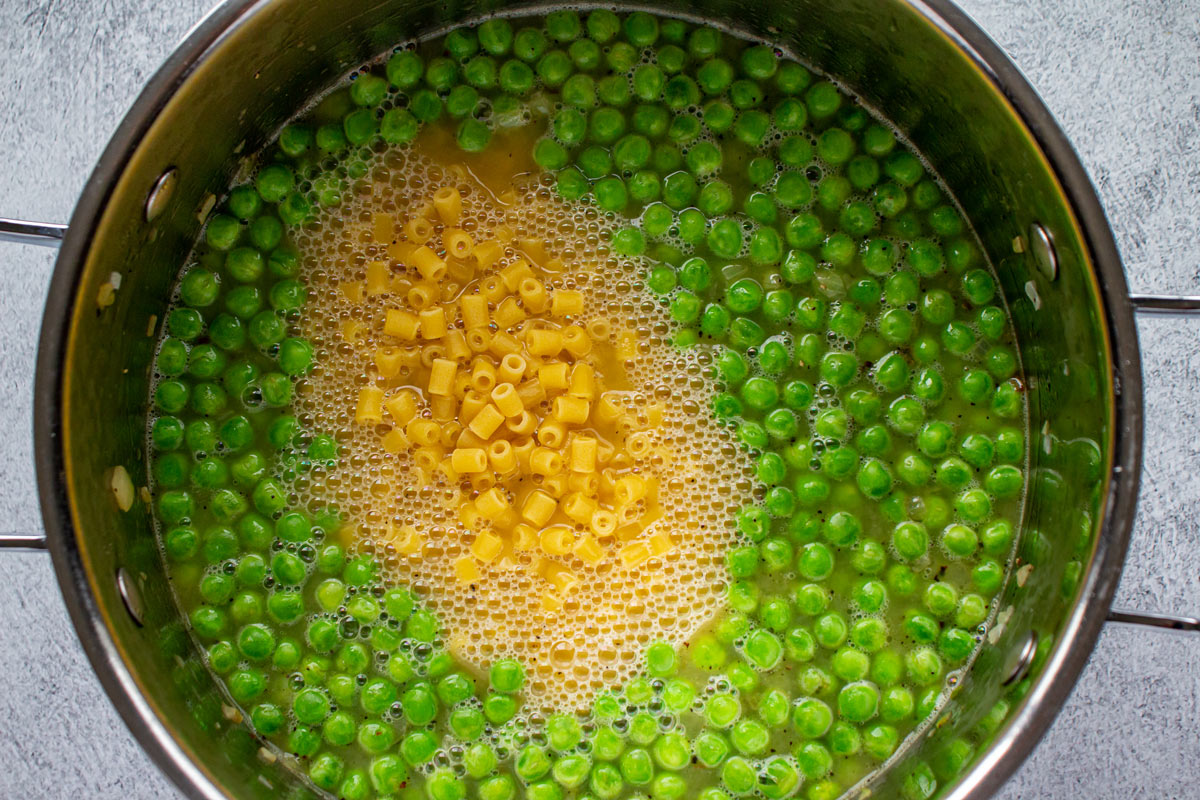 Peas and uncooked ditalini pasta in broth in a metal pot.