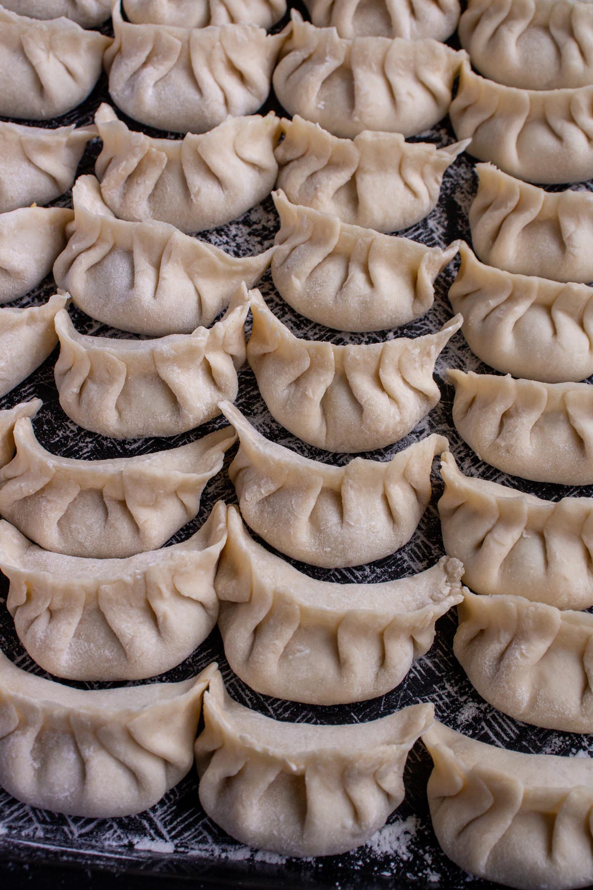 Four rows of assembled but uncooked Chinese dumplings with pleated edges on a black surface.