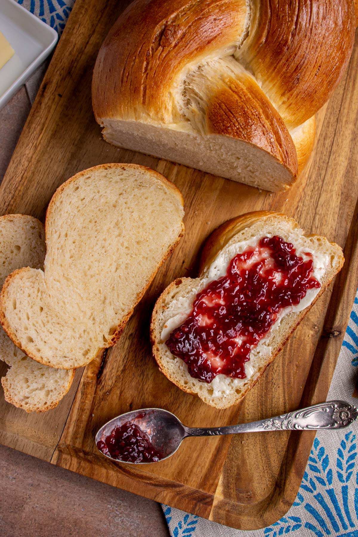Slices of white braided bread on a wooden board, one topped with butter and raspberry jam.