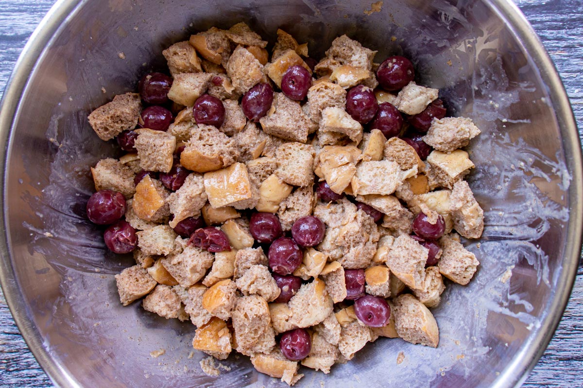 A mixture of soaked bread cubes and cherries in a metal mixing bowl.