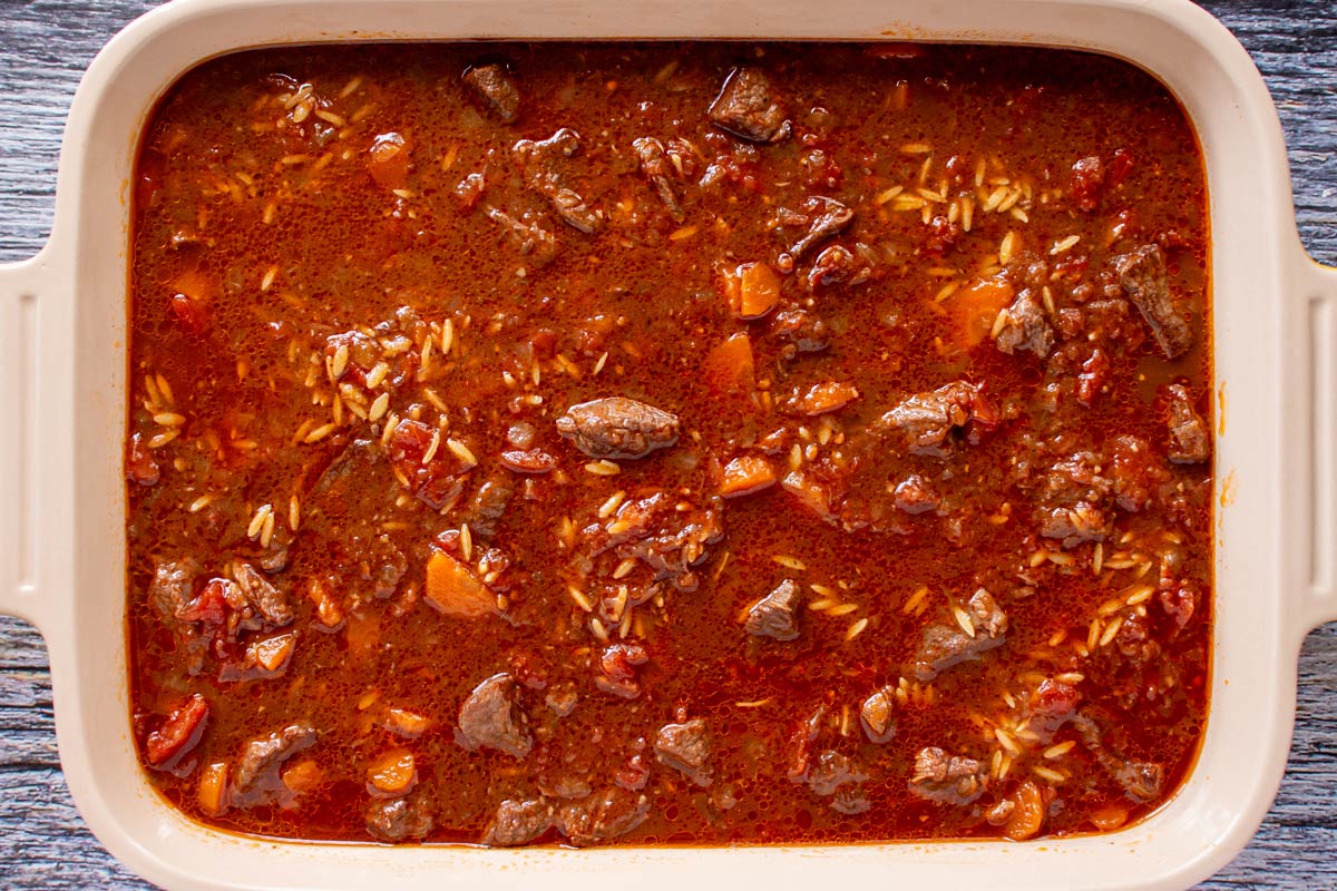A beef stew mixture in a rectangular baking dish with some orzo visible through the liquid.