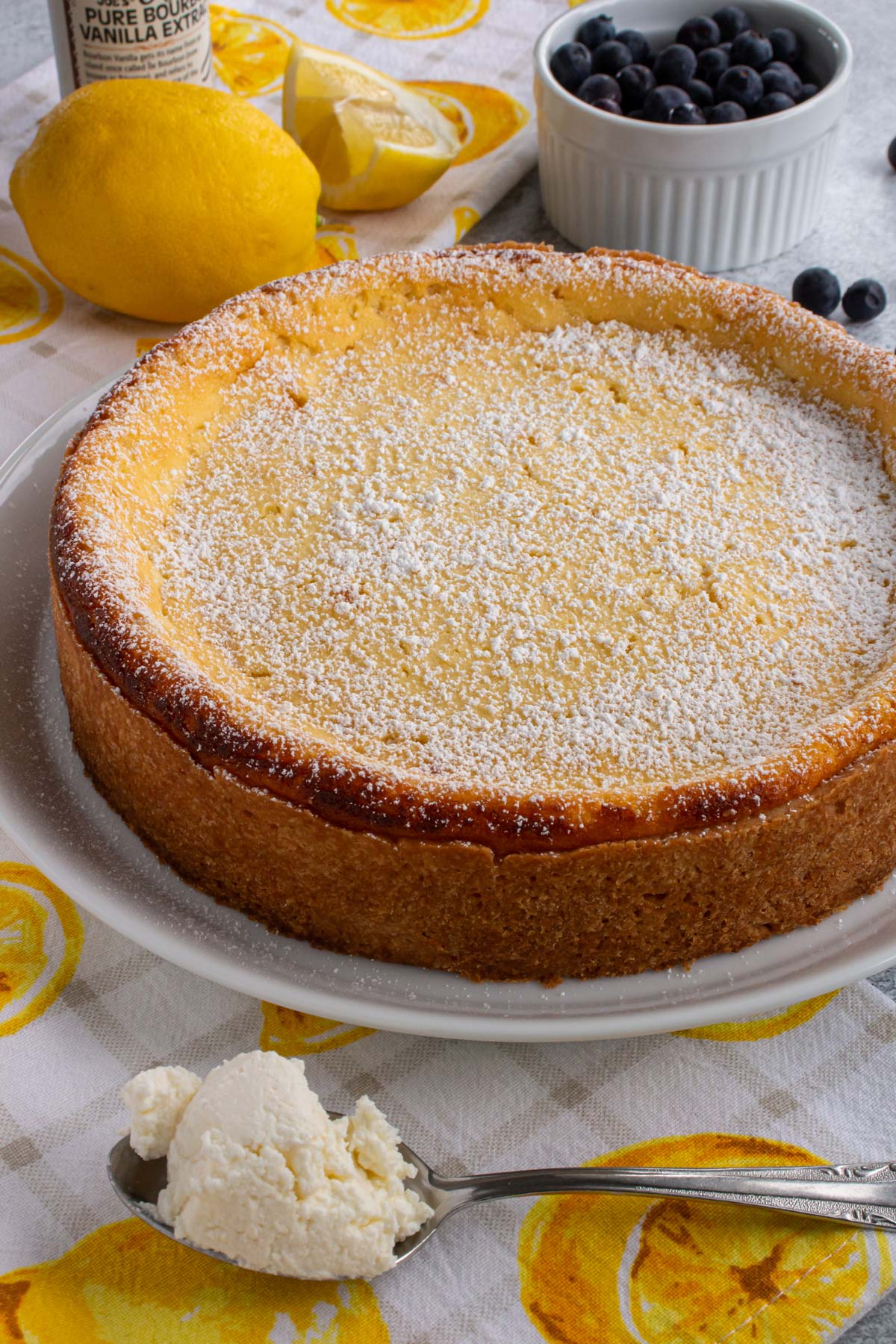 A German cheesecake topped with powdered sugar with lemons and blueberries in the background.