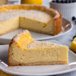 A slice of käsekuchen on a plate with the remaining cheesecake in the background.