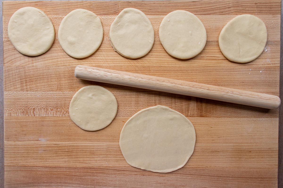 Rounds of dough being rolled out thinner into larger circles on a wooden board.