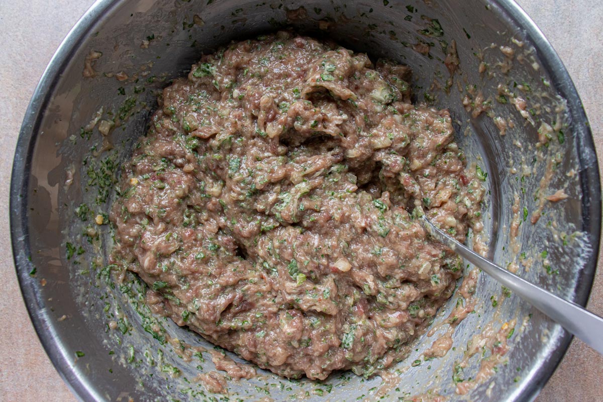 A raw meat mixture with herbs in a metal bowl with a spoon.