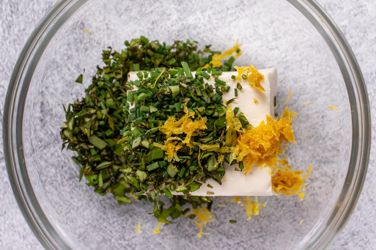 A block of cream cheese, chopped herbs, and lemon zest in a glass bowl.