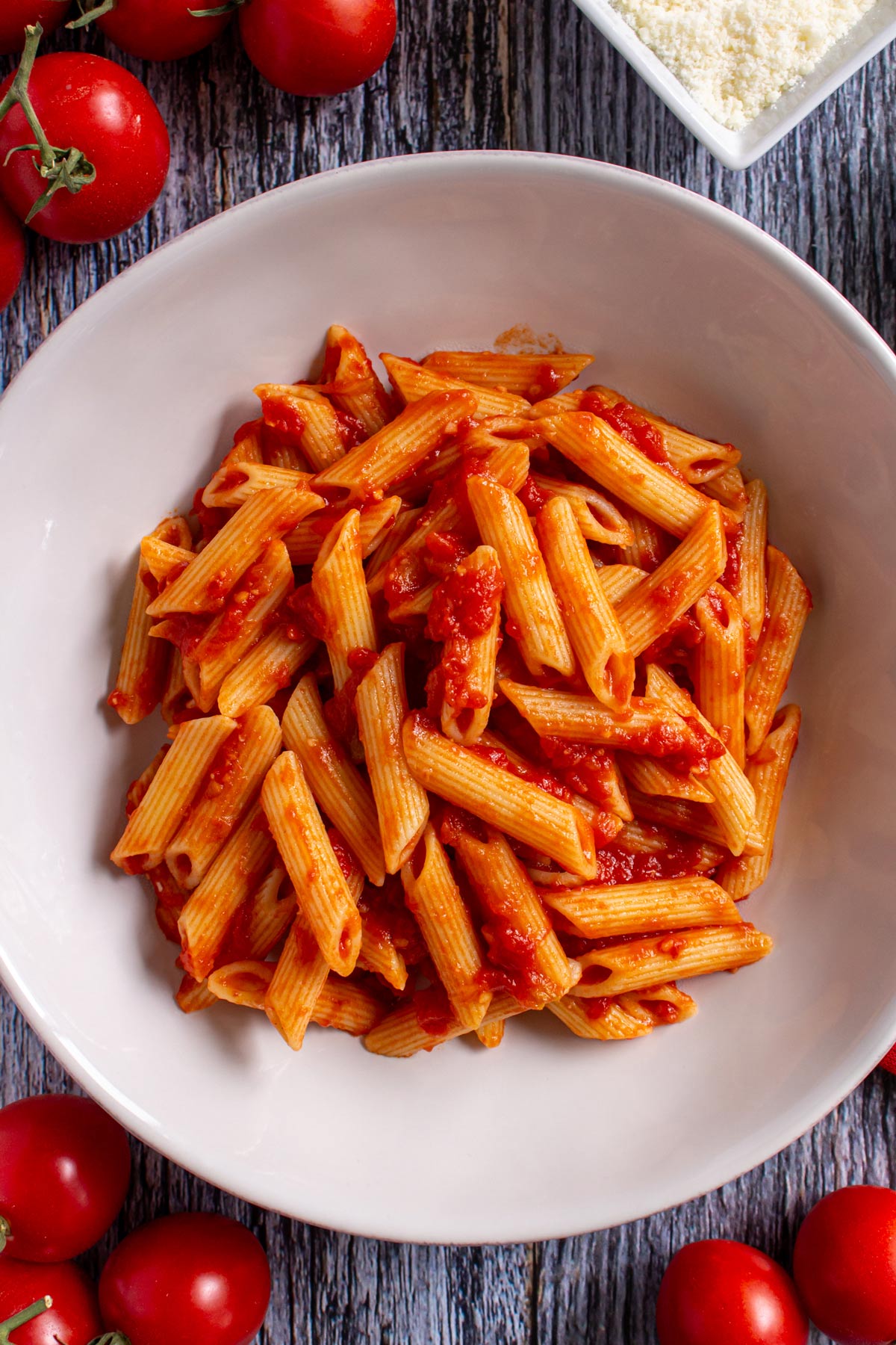 Penne arrabbiata in a wide pasta bowl surrounded by small tomatoes on a wooden background.