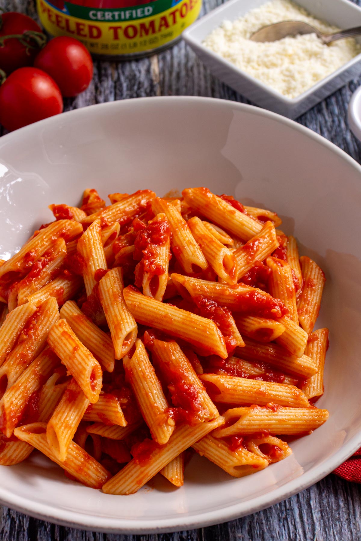 Penne all' arrabbiata in a pasta bowl with tomatoes and grated cheese in the background.