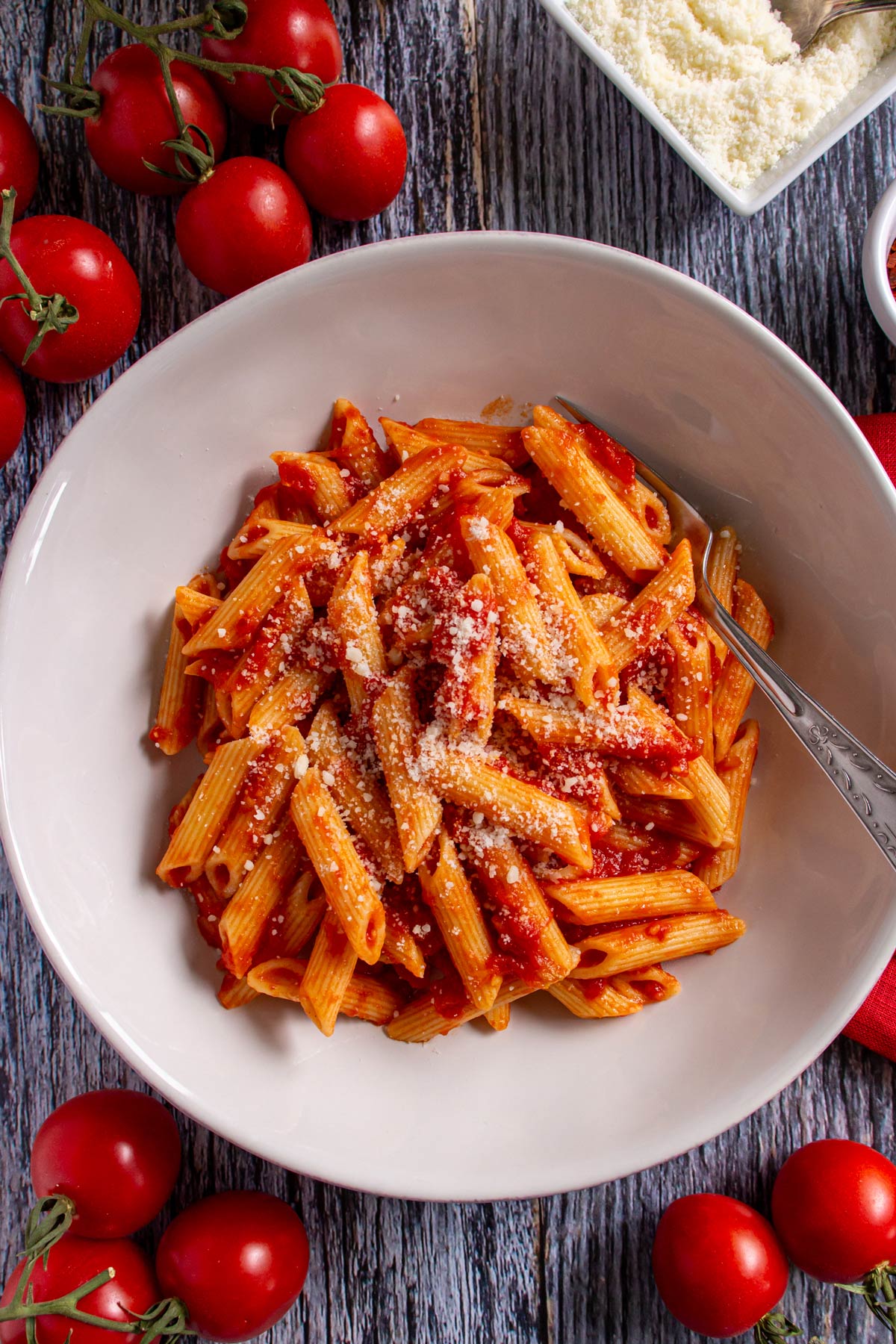Penne all' arrabbiata topped with cheese in a wide pasta bowl surrounded by small tomatoes.