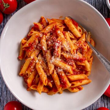 Penne all' arrabbiata topped with cheese in a wide pasta bowl surrounded by small tomatoes.