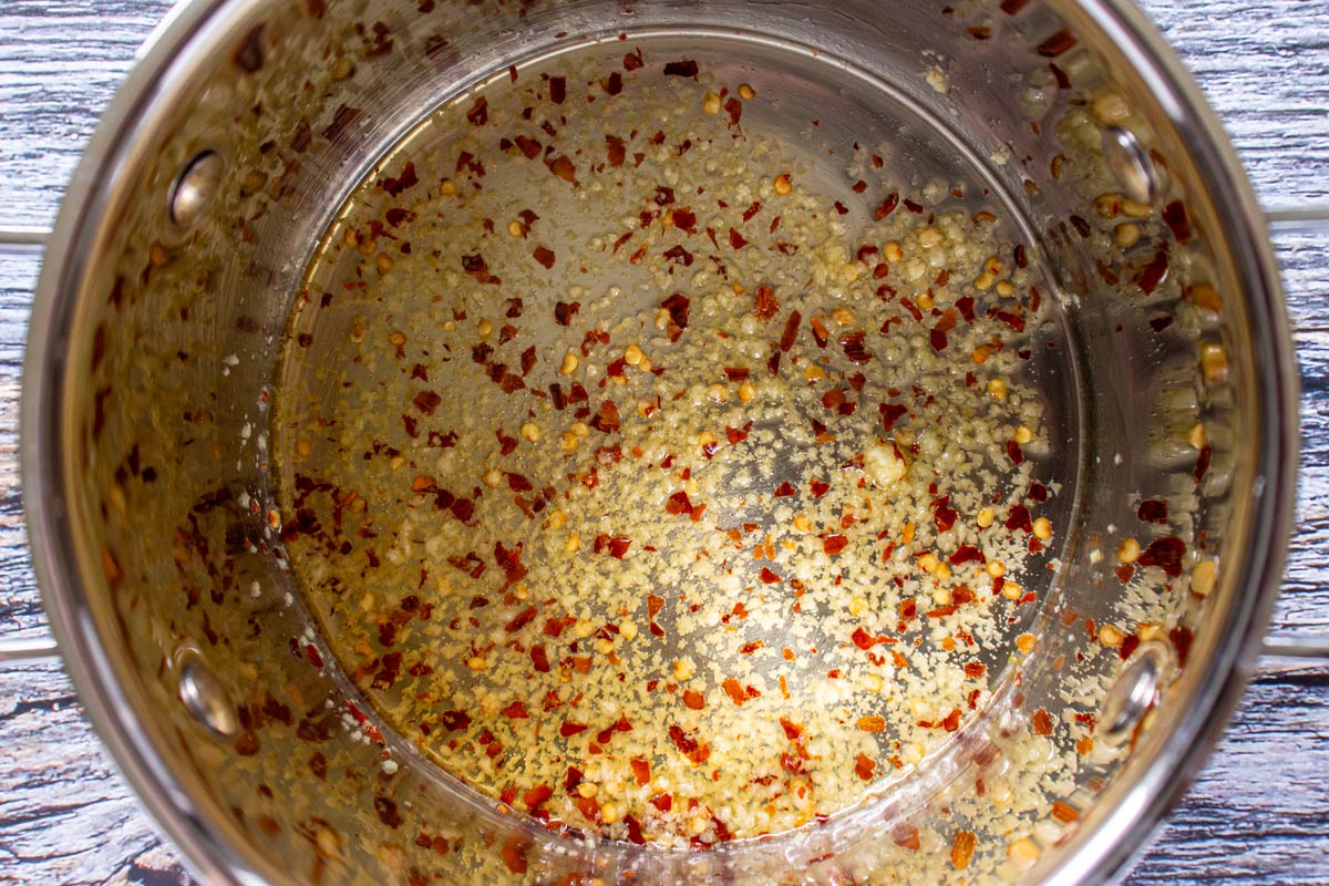 Crushed garlic and chili flakes cooking in olive oil in a metal pot.