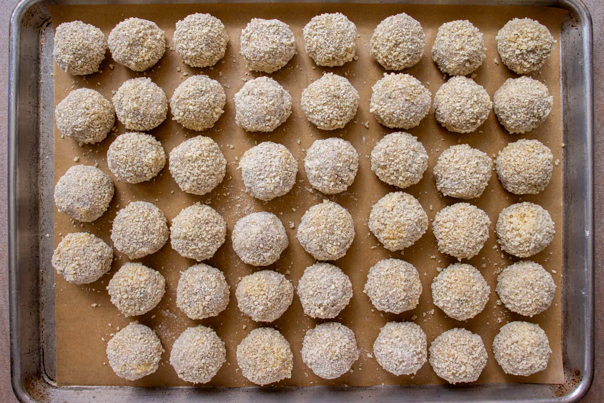Breaded but uncooked balls arranged in rows on a parchment paper lined baking sheet.