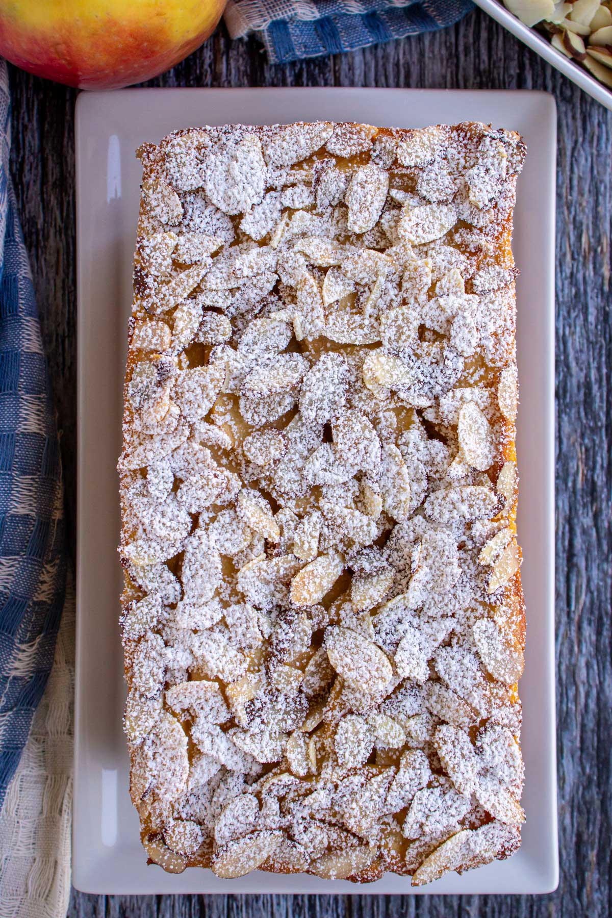 A rectangular invisible apple cake topped with powdered sugar dusted sliced almonds.