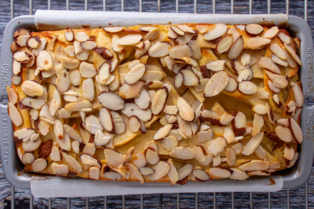 A baked invisible apple cake topped with almonds in a loaf pan cooling on a rack.
