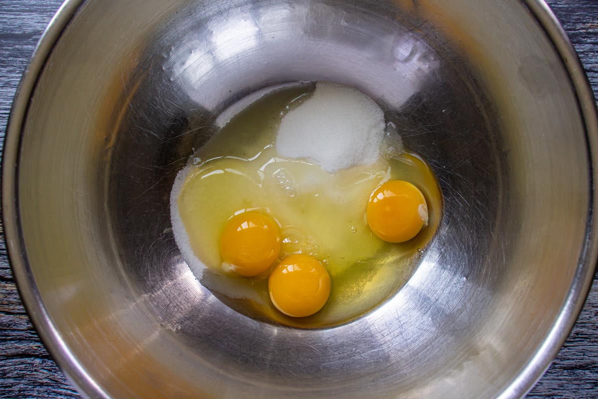 White sugar and 3 cracked eggs in a metal mixing bowl.