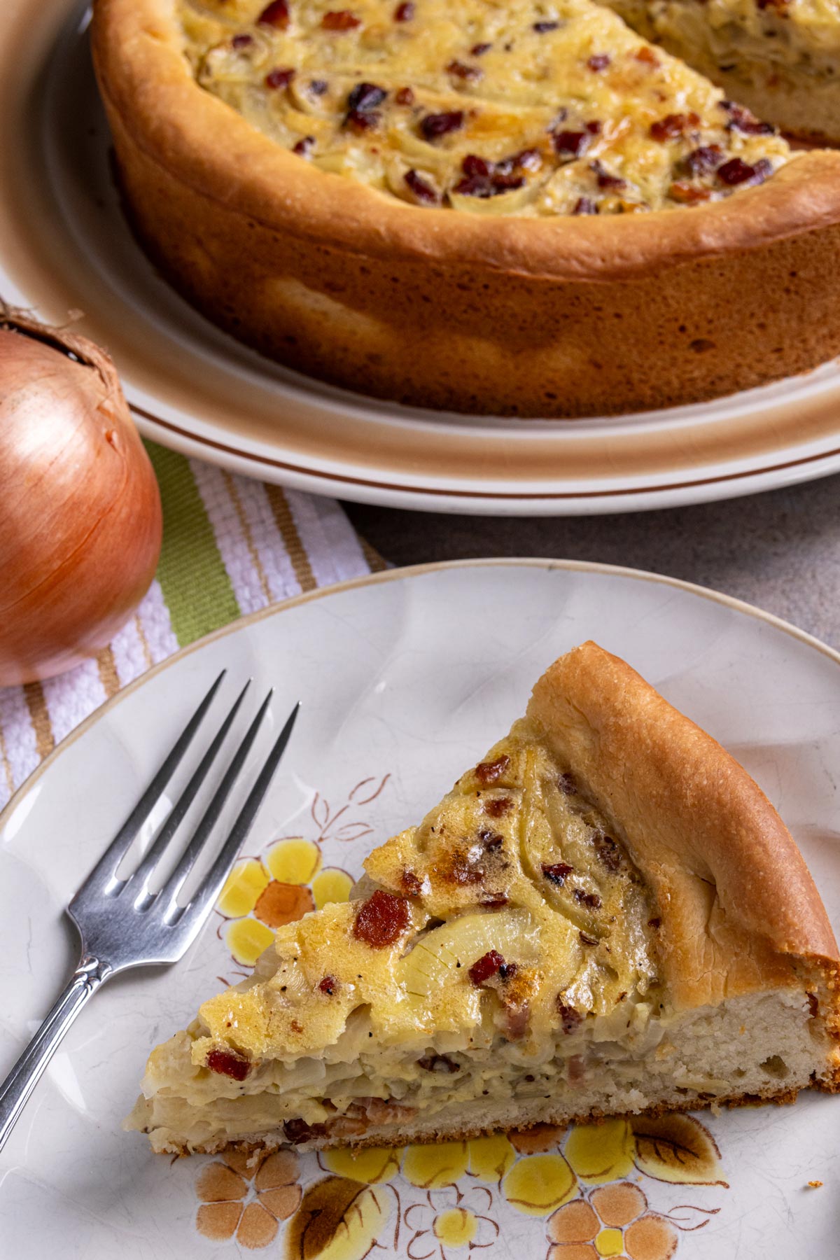 A slice of zwiebelkuchen on a plate with a fork, with the remaining onion pie behind it.