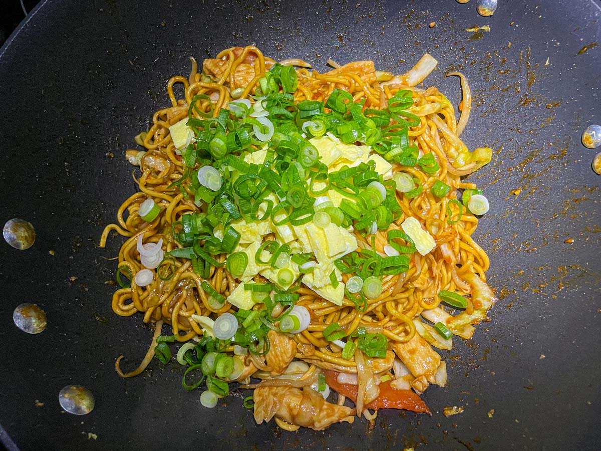 Adding chopped omelet and scallions to a wok filled with stir-fried noodles.