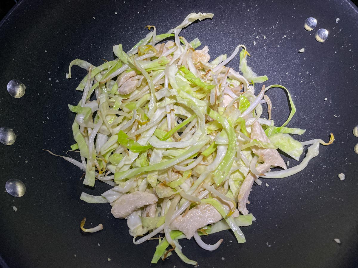 A mixture of sliced chicken, bean sprouts and cabbage cooking in a wok.
