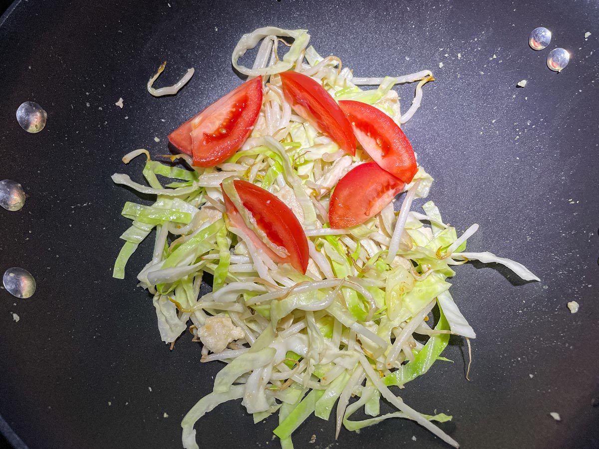 Adding wedges of tomato to a wok with bean sprouts, cabbage, and chicken.