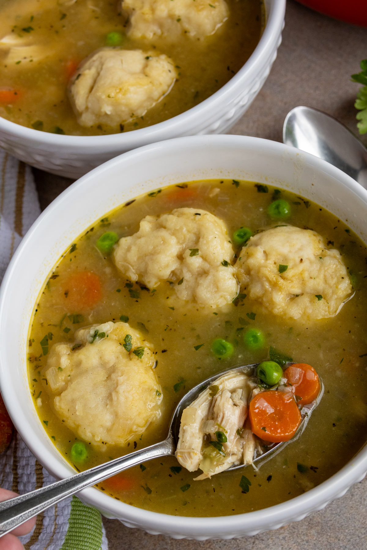 A spoon lifting out chicken and vegetables out of a white bowl of chicken and dumplings.