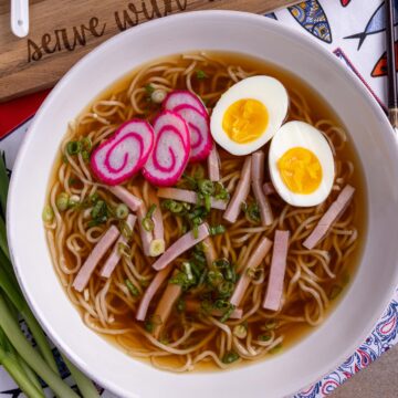 A bowl of saimin garnished with ham, scallions, fish cakes, and a halved boiled egg.